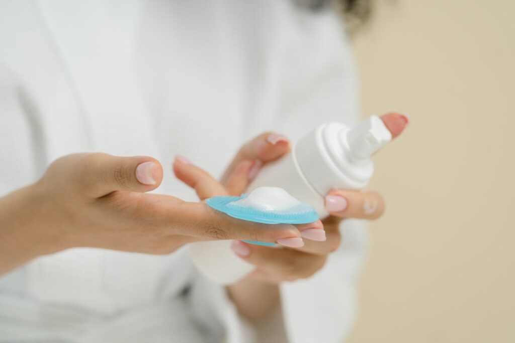 Close-up of hands holding a bottle dispensing skincare foam onto a cleansing pad, showcasing manicured nails.